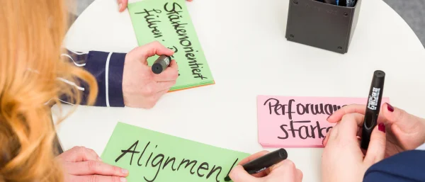 Drei Führungskräfte stehen um einen Tisch und beschreiben Karten mit den Worten " Alignment", "stärkenorientiert führen" und "performance stärken"