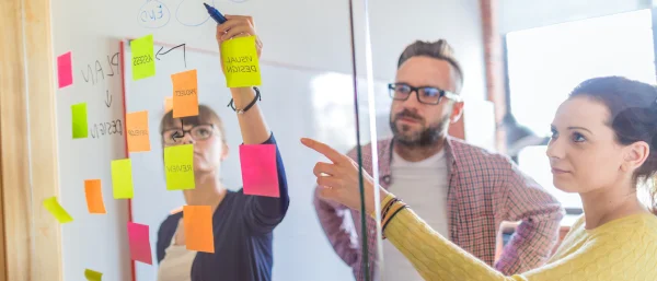 Drei Personen stehen vor einer Glaswand mit Post It's und schreiben darauf.