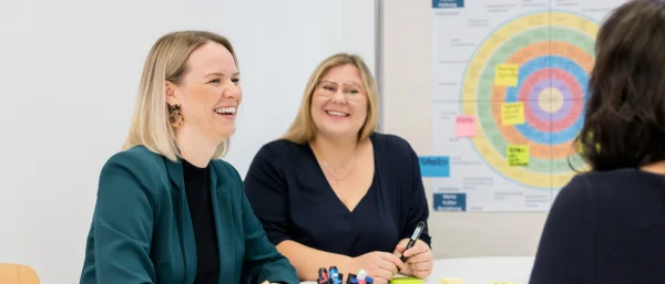 Jana Schmid und Bettina Golba - Trainerinnen bei Carpe Verba lachen freundlich und sind im Austausch über integrale Organisationsentwicklung. Im Hintergrund ist ein Plakat mit einem bunten Kreis.