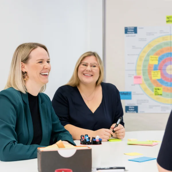 Jana Schmid und Bettina Golba - Trainerinnen bei Carpe Verba lachen freundlich und sind im Austausch über integrale Organisationsentwicklung. Im Hintergrund ist ein Plakat mit einem bunten Kreis.
