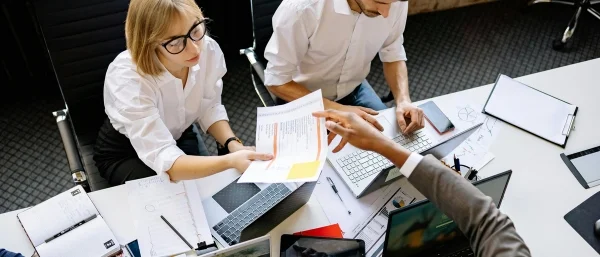 Drei Personen sitzen gemeinsam am Tisch und betrachten in Zusammenarbeit ein Dokument im Ausdruck einer gemeinsamen Teamkultur