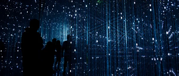 eine Gruppe von Menschen steht in einem dunklen Raum, in dem viele kleine Lichter zu sehen sind. Die Personen nicht aufgrund der Dunkelheit nicht kenntlich. Es wirkt zukunftsorientiert, technologisiert.