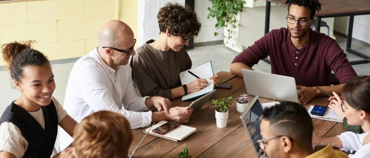Sieben Mitarbeitende sitzen in Kooperation im Stil der Arbeitswelt 4.0 um einen Tisch herum und freuen sich über die gute Zusammenarbeit.