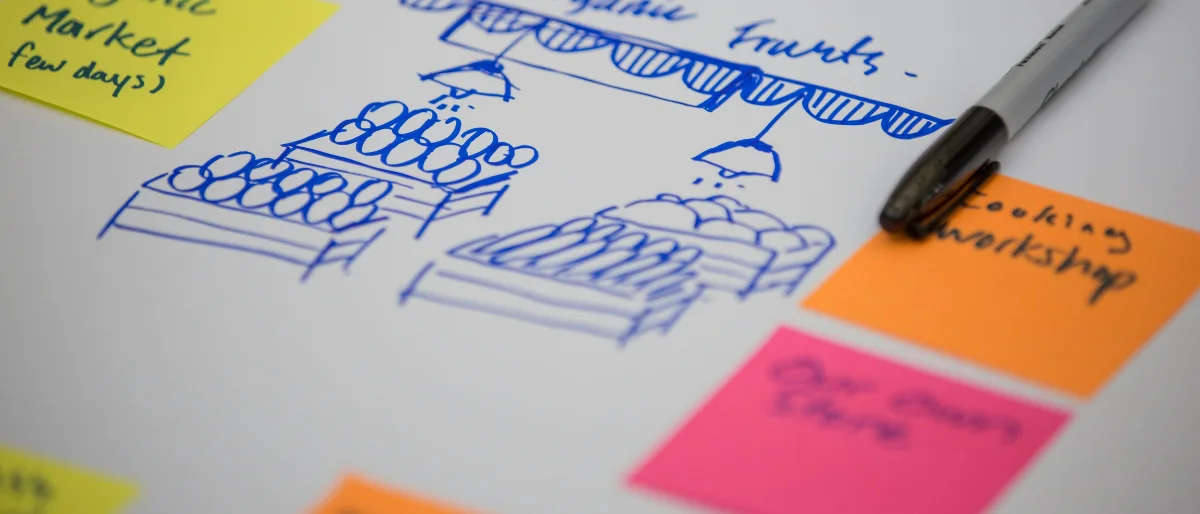 Es ist eine prototypen Skizze eines Marktstandes zu sehen, rechts und links davon kleben bunte Sticky Notes und es liegt ein Stift. Alles auf einer weißen Oberfläche