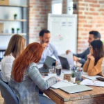 5 Personen sitzen konzentriert um einen Holztisch, im Hintergrund steht eine Flipchart, was auf einen Workshop hindeutet. Die Teilnehmenden wirken auch durch ihre Laptops konzentriert und arbeitsam.