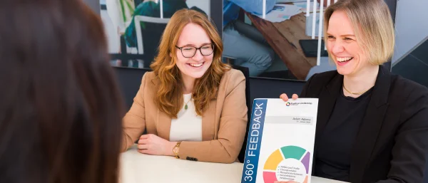 Auf dem Bild sind zwei lächelnde Trainerinnen von Carpe Verba zu sehen. Eine von ihnen hält ein Mappe zum Thema 360-Grad-Feedback für die Organisationsdiagnostik mit PERMA-Lead.