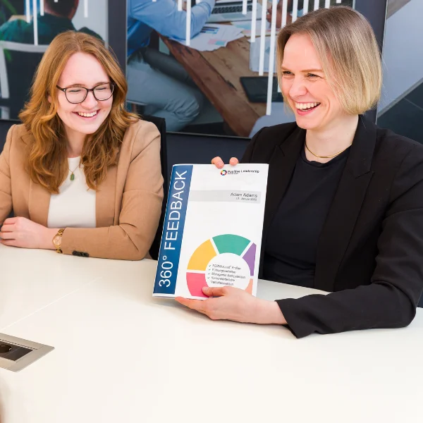 Auf dem Bild sind zwei lächelnde Trainerinnen von Carpe Verba zu sehen. Eine von ihnen hält ein Mappe zum Thema 360-Grad-Feedback für die Organisationsdiagnostik mit PERMA-Lead.