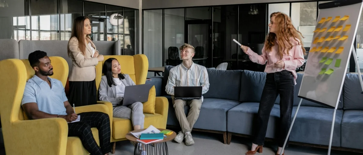 Fünf junge Erwachsene in moderner Businesskleidung befinden sich in einem offenen, kreativ gestalteten Meetingraum. Zwei Personen sitzen in leuchtend gelben Sesseln, zwei weitere auf einem Sofa – alle wirken aufmerksam und interessiert. Eine Frau mit langem, lockigem Haar steht neben einem Flipchart, das mit bunten Post-its bestückt ist, und spricht engagiert zur Gruppe. Laptops und Notizblöcke deuten auf eine kollaborative Arbeitsatmosphäre hin. Das Setting strahlt Teamwork, Innovation und eine moderne Arbeitskultur aus.