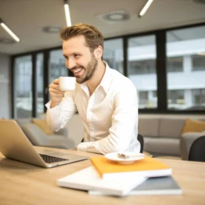 Ein Mann in weißem Hemd sitzt lächelnd vor einem Laptop und trinkt Kaffee. Das Bild steht symbolisch für "Stresskompetenz: souverän führen".