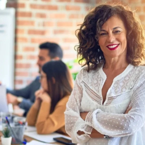 In einem modernen Büro mit Backsteinwand steht eine selbstbewusste Frau mit lockigem Haar und weißer Bluse im Vordergrund, lächelnd und mit verschränkten Armen. Im Hintergrund arbeitet ein diverses Team konzentriert an einem großen Tisch – manche tippen auf Laptops, andere diskutieren miteinander. Auf einem Whiteboard sind Notizen und Symbole zu erkennen. Die Szene vermittelt eine Atmosphäre von Leadership, Vertrauen und produktiver Zusammenarbeit.