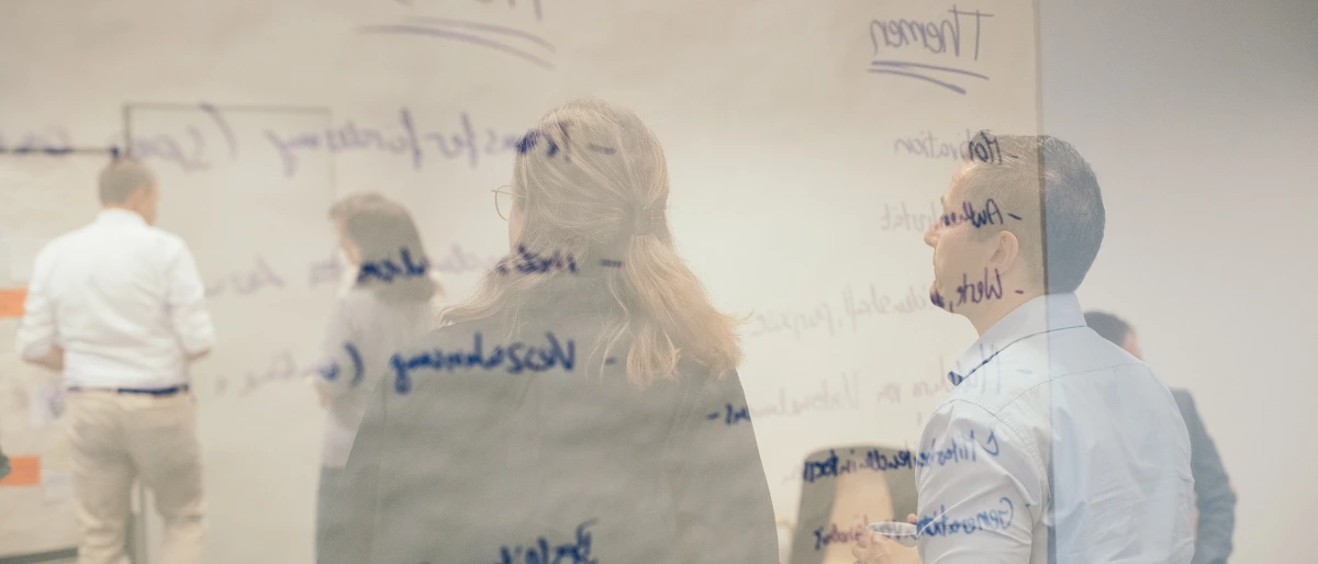 Im Vordergrund des Bildes sieht man einen beschrifteten Flip-Chart und im Hintergrund ein Team, das gerade einen Teamworkshop durchführt.